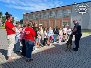 Na placu szkoły policjant z psem służbowym oraz grupa dzieci wraz z opiekunami