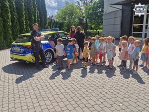 Grupa dzieci na zewnątrz przedszkola wraz z policjantem i opiekunkami obok radiowozu