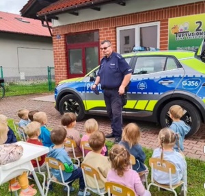 Policjant na tle radiowozu prowadzący pogadankę dla przedszkolaków siedzących na krzesełkach przed budynkiem