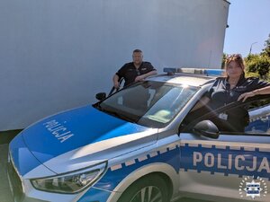 Dwójka policjantów po obu stronach radiowozu na tle ściany budynku