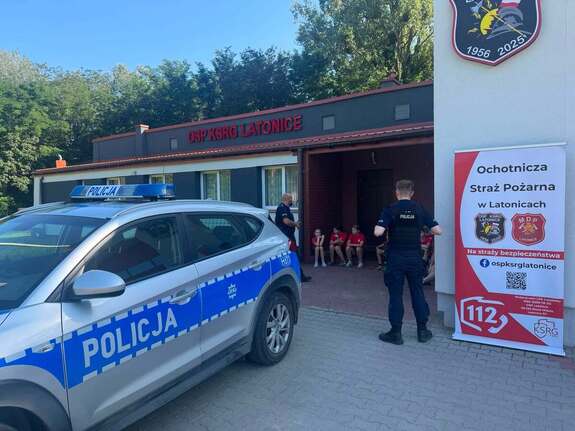 Radiowóz przed strażnicą, dwaj policjanci a wewnątrz grupa młodzieży w czerwonych koszulkach