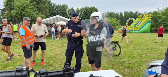 Na pikniku na tle zieleni młody chłopak ubrany w kamizelkę ochronną kask i tarczę, obok policjant, w tle grupy ludzi