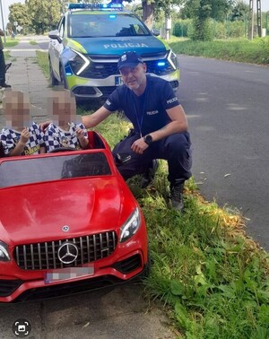 Policjant na tle radiowozu przy chodniku, na którym znajduje się dwójka dzieci w samochodziku zabawkowym