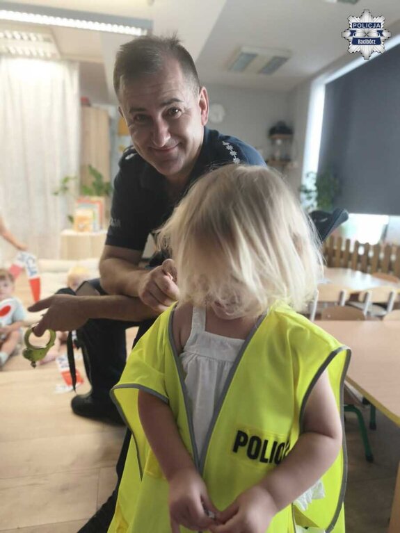 Uśmiechnięty policjant pochylający się nad dzieckiem ubranym w kamizelkę odblaskową z napisem Policja