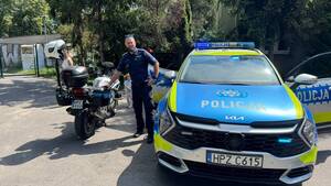 Na dziedzińcu, na tle zieleni pomiędzy radiowozem a motocyklem policyjnym policjant z brodą
