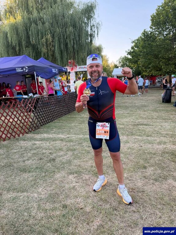 mężczyzna w stroju sportowym w prawej ręce trzyma zdobyty medal, lewa ręka zaciśnięta w pięść uniesiona na wysokości głowy.