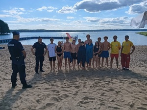 na brzegu jeziora, przed grupą młodzieży i dwóch ratowników wodnych stoi policjant w mundurze