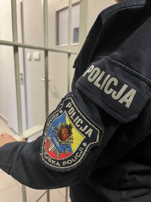 na tle metalowych krat widnieje fragment ramienia umundurowanego policjanta, na ramieniu widnieje biały napis POLICJA