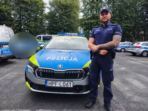 policjant w umundurowaniu służbowym stoi przed oznakowanym radiowozem z napisem POLICJA