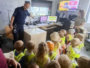 umundurowany funkcjonariusz Policji prezentuje gromadce dzieci stanowisko dyżurnego jednostki Policji