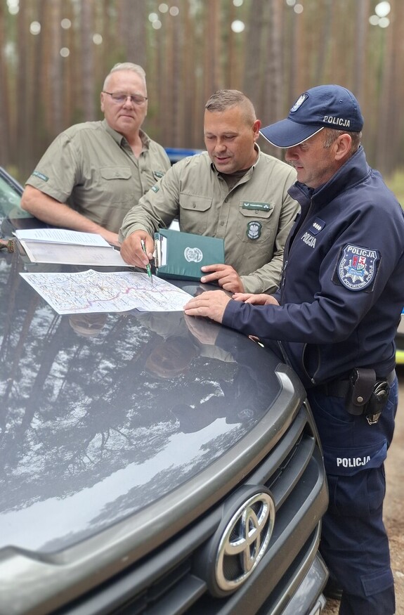 trzej mężczyźni stoją przy  masce samochodu, na masce rozłożona jest mapa, dwóch mężczyzn w mundurze straży leśnej a jeden mężczyzna w umundurowaniu policyjnym.