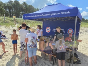 umundurowana policjantka wraz z kobietą w białej koszulce rozmawiają z dziećmi siedzącymi przy stole, który znajduje się pod namiotem z napisem Powiatowa Stacja Sanitarno-Epidemiologiczna w Sławnie