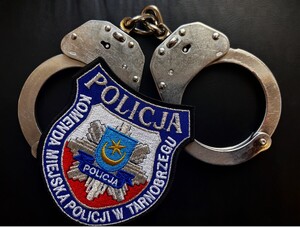 na czarnym tle ułożone są kajdanki policyjne, na których leży naszywka policyjna z napisem Komenda Miejska Policji w Tarnobrzegu