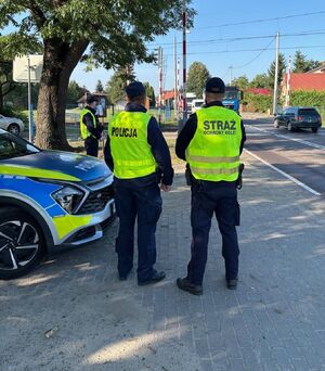 dwóch umundurowanych policjantów w kamizelkach odblaskowych z napisem Policja, stoją przed oznakowanym radiowozem na tle przejazdu kolejowego