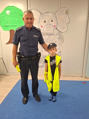 umundurowany policjant stoi z małym chłopcem, który ma na głowie policyjną czapkę z daszkiem i ubrany jest w kamizelkę odblaskową