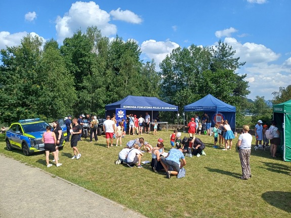 Piknik na otwartej zielonej przestrzeni, centralnie namioty wystawiennicze, obok dzieci i radiowóz