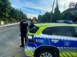 dwóch umundurowanych policjantów stoją przed oznakowanym radiowozem w rejonie zejścia pieszego przy szkole