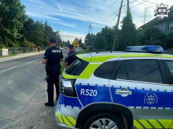 dwóch umundurowanych policjantów stoją przed oznakowanym radiowozem w rejonie zejścia pieszego przy szkole