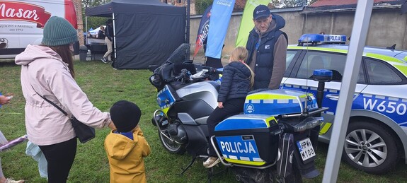 Piknik na terenie zielonym w tle namioty wystawiennicze na pierwszym planie dziecko na motocyklu policyjnym wraz z opiekunem obok radiowóz
