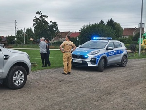 Na żużlowej drodze osiedla radiowozów, samochód terenowy, oraz policjant i cywil wskazujący ręką
