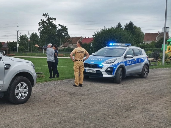 Na żużlowej drodze osiedla radiowozów, samochód terenowy, oraz policjant i cywil wskazujący ręką
