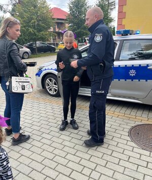 Policjant wręczający na chodniku ulotkę dziewczynce, obok kobieta w tle radiowóz