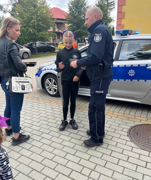 Policjant wręczający na chodniku ulotkę dziewczynce, obok kobieta w tle radiowóz
