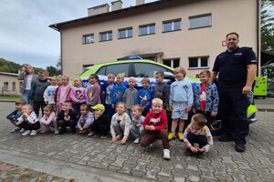 Dzieci pozujące do zdjęcia grupowego wraz z policjantem i wychowawczynią na tle radiowozu i budynku