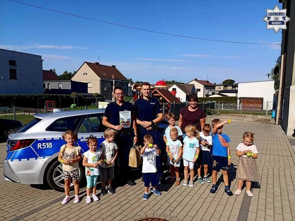Grupa przedszkolaków pozująca wraz z dwoma policjantami i wychowawczynią do zdjęcia na tle radiowozu