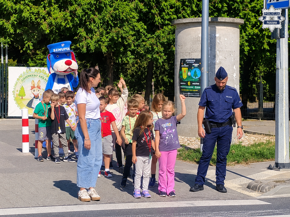 Przed przejściem dla pieszych policjant z grupką dzieci oraz wychowawczynią za nimi maskotka Sznupek