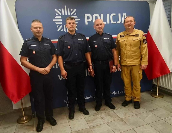 Na tle policyjnej ścianki i flag państwowych: trójka policjantów i strażak