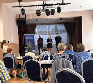 Dwaj policjanci oraz kobieta prowadzący spotkanie dla zgromadzonych seniorów