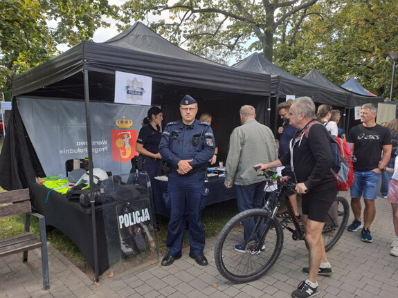 Policjant przed namiotem wystawienniczym, obok senior z rowerem, w tle inni seniorzy