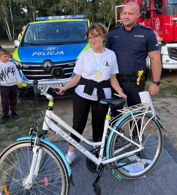 Uśmiechnięta kobieta z rowerem, obok policjant na tle zieleni radiowozu i wozu bojowego straży pożarnej