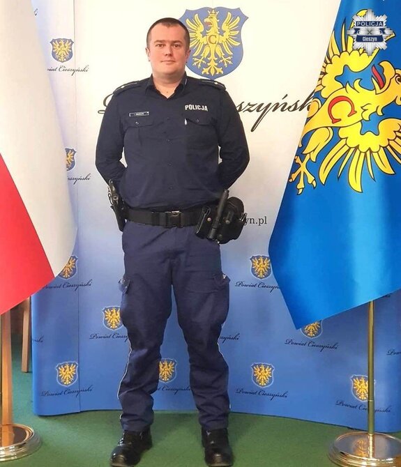 Policjant pozujący do zdjęcia na tle flag i ścianki powiatu cieszyńskiego