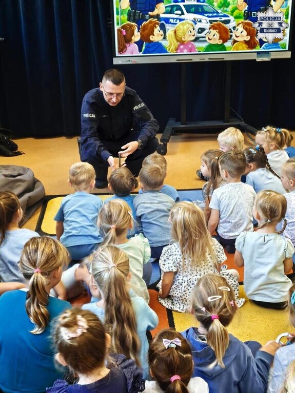 Policjant prowadzący pogadankę dla dzieci siedzących na wykładzinie