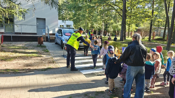 Na terenie zielonym przedszkola grupa maluchów, policjantka oraz wychowawczyni, w tle radiowóz
