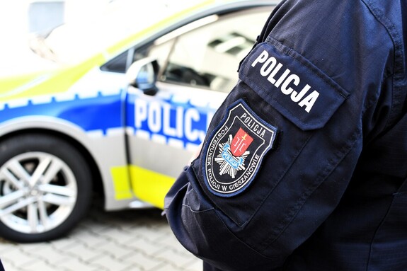 Ramię policjanta w kurtce służbowej z napisem Posterunek Policji w Cieszanowie, w tle radiowóz