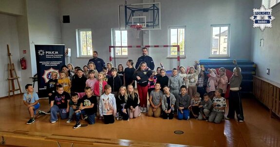 Dwaj policjanci pozujący do zdjęcia z grupą dzieci wewnątrz sali gimnastycznej