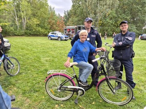 W scenerii zielonego pikniku dwaj policjanci i uśmiechnięta kobieta z rowerem