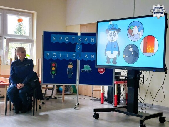 Policjantka w sali lekcyjnej gdzie na tablicach wyświetlane są rysunki z napisem "Spotkanie z policjantem"