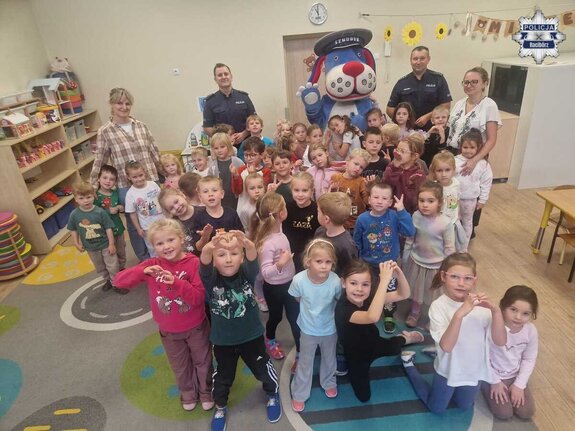 Przedszkolaki pozujące do zdjęcia w sali wraz z wychowawczyniami, dwoma policjantami oraz maskotką "Sznupek"