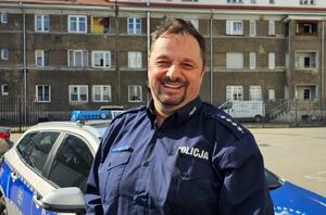 Uśmiechnięty policjant na tle radiowozu i budynku wielorodzinnego