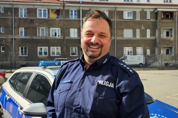 Uśmiechnięty policjant na tle radiowozu i budynku wielorodzinnego