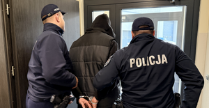 Dwaj policjanci wyprowadzający z budynku mężczyznę ze skutymi kajdankami rękami i w bluzie z nasuniętym na głowę kapturze