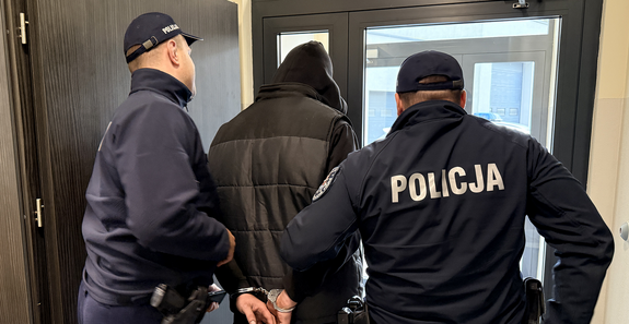 Dwaj policjanci wyprowadzający z budynku mężczyznę ze skutymi kajdankami rękami i w bluzie z nasuniętym na głowę kapturze