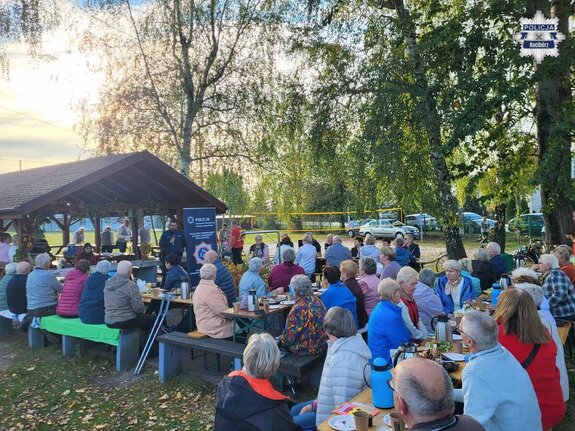 Piknik na otwartym zielonym terenie, przy stołach seniorzy, w głębi widoczny policjant prowadzący spotkanie
