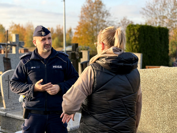 Zdjęcie przedstawiające policjanta na tle cmentarza prowadzącego rozmowę z kobietą,